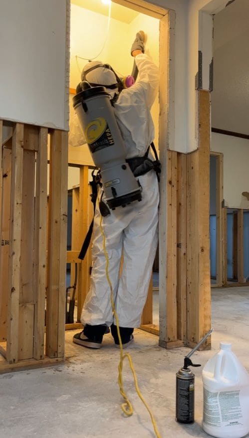 AmeriTribe technician in a protective suit and respirator cleaning exposed wall framing with a HEPA vacuum